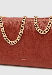 Rdeča usnjena torbica z teksturo, zlato verižico in izbočenim logotipom. Ima pravokotno obliko in gladko površino.