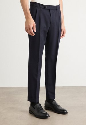 Navy pleated trousers with a tailored fit, featuring a button closure, cuffed hems, and smooth fabric texture, paired with black loafers.