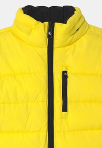 Žlutá zateplená bunda s vysokým límečkem, černým zipem a jemně prošívanou texturou. Obsahuje přední kapsu na zip.