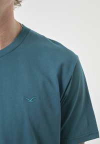 Türkisfarbene Baumwoll-T-Shirt mit Rundhalsausschnitt, das ein kleines blau gesticktes Vogel-Logo auf der linken Brustseite zeigt. Glatte Textur, lässiges Design.