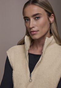 Beżowa kamizelka polarowa z wysokim kołnierzem i srebrnym zamkiem błyskawicznym. Kamizelka ma miękką fakturę i jest noszona na czarnym topie z długim rękawem.