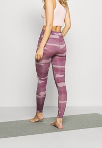 Fioletowe legginsy w kamuflażu o wysokim stanie, miękkiej strukturze i oddychającym materiale. Modelka stoi boso na szarej macie.