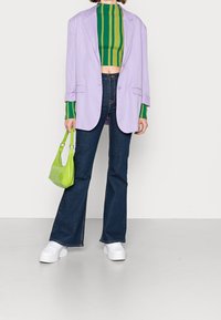 Blazer oversize viola chiaro, top corto a righe verdi e gialle, jeans a zampa d'elefante blu scuro, sneakers bianche e una borsa verde piccola.