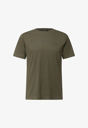 Mężczyzna z krótkimi włosami i brodą nosi oliwkową koszulkę z napisem "Zakład Przemyślanych Wyrobów Bezkonkurencyjny Komfort" z przodu.