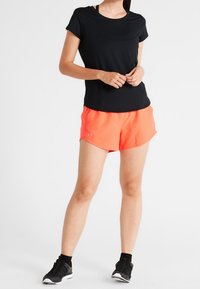 Czarna, krótko rękawowa koszulka sportowa w połączeniu z jasnopomarańczowymi szortami z siateczki. Model nosi czarne buty biegowe i skarpetki ankle.