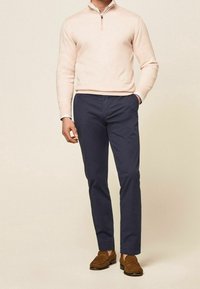 Světle růžový svetr na zip s výstřihem u krku v kombinaci s tmavě modrými kalhotami a hnědými semišovými mokasínami. Pod svetrem jsou vidět manžety bílého košile.