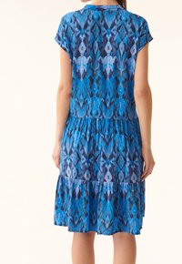 Vestido azul con estampado, de mangas cortas, que presenta una falda en capas y una mezcla de diseños geométricos en diversas tonalidades de azul y púrpura.