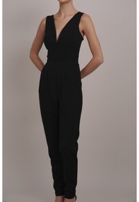 Mono negro con escote en V profundo, diseño sin mangas, corte ajustado, pliegues en la cintura y piernas entalladas, fabricado en tejido suave.