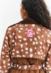 Brązowy płaszcz w wzory z imitacją futra w białe plamki, z paskiem w talii oraz różową metką z grafiką grzyba.