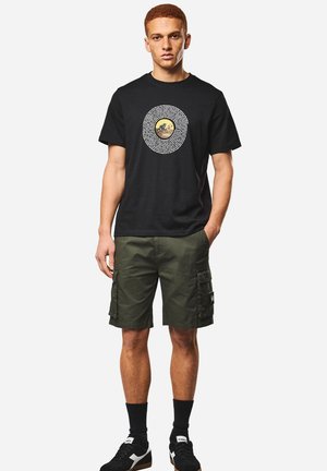 Junger Mann, der ein schwarzes T-Shirt mit Grafik, olivgrüne Cargoshorts, schwarze Socken und schwarz-weiße Turnschuhe trägt, steht mit einer Hand in der Tasche.