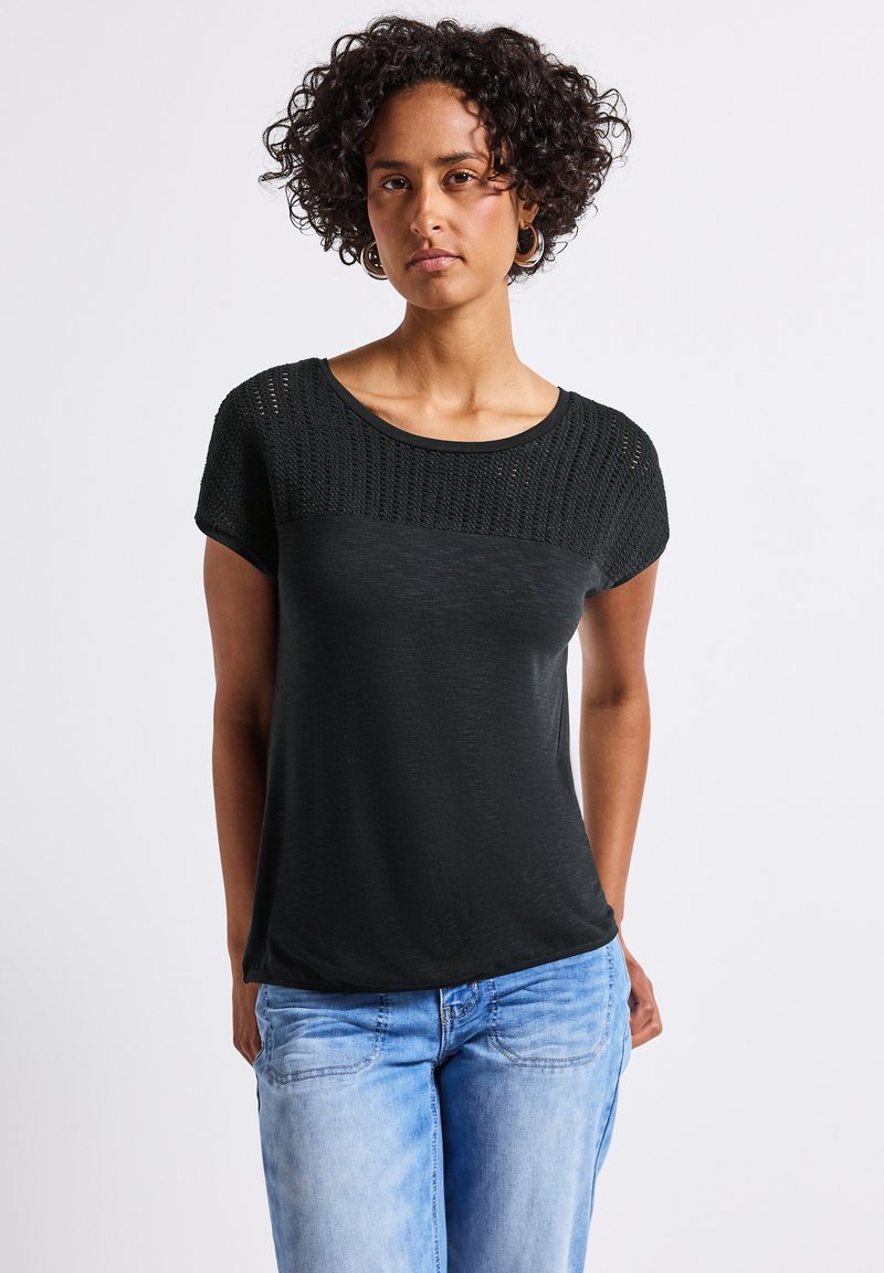 Haut noir à manches courtes avec un design en maille texturée sur les épaules et un corps lisse, porté avec un jean bleu clair.