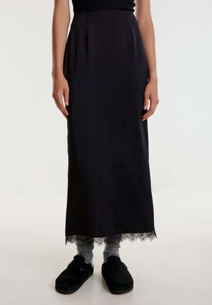 Jupe maxi noire en tissu lisse, avec un bord en dentelle à l'ourlet. Le modèle porte des chaussons foncés sur des chaussettes grises.