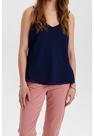 Jeune femme aux longs cheveux portant un haut sans manches bleu marine et un pantalon rose clair, debout devant un fond clair uni.