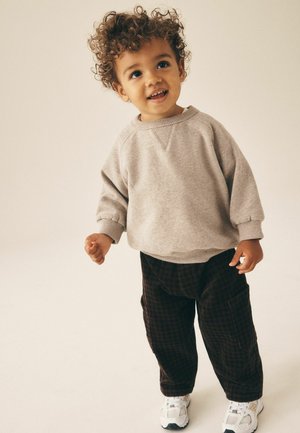 Småbarn med krøllet hår iført beige sweatshirt, mørke ternede bukser og hvide sneakers, står og kigger op med et smil.