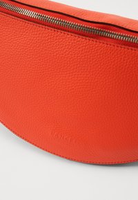Pomarańczowa skórzana kopertówka o chropowatej fakturze, z metalowym suwakiem u góry i wytłoczonym logo "LANCEL" z przodu.