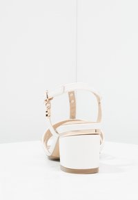 Valkoiset korkokengät, joissa on leveä korko ja nilkkahihna, sisältäen kiiltävän kullanvärisen yksityiskohdan perustassa ja sileän nahkapinnan.