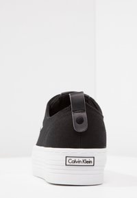 Sneaker nera in tela con suola piatta bianca, dotata di un tassello in pelle nera e un'etichetta logo sul tallone. Design e texture puliti.