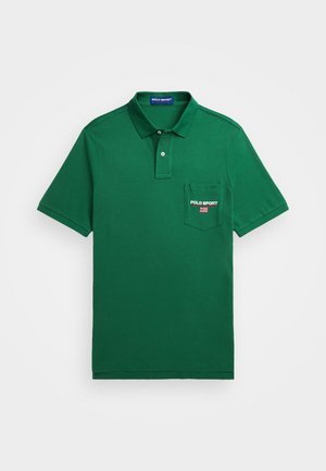 Osoba nosząca zieloną koszulkę Polo Sport z naszywką z amerykańską flagą, paskiem w paski, dżinsowymi szortami poplamionymi farbą oraz niebieskim zegarkiem na lewej ręce.