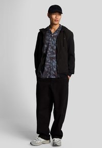 Giacca nera con zip sopra una maglietta blu scuro a tie-dye, pantaloni neri e scarpe da corsa grigie con accenti bianchi. Un cappellino grigio completa l'outfit.