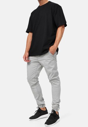 Mand iført en ensfarvet sort kortærmet t-shirt, lysegrå slim joggerbukser og sorte sneakers med hvide såler, stående med den ene hånd i lommen.