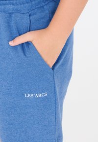 Siniset puuvillahousut pehmeällä tekstuurilla, joissa on sivutasku ja pieni valkoinen "LES' ARCS" -logo helmassa.