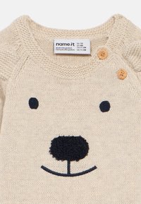 Pull en maille beige avec un motif de visage d'ours, broderie marine, détail de bouton en bois à l'épaule et texture douce.