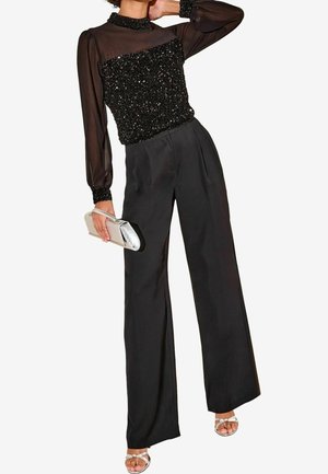 Femme portant un haut noir à sequins avec manches longues transparentes et col montant, pantalon noir à jambes larges, tenant une pochette argentée et portant des sandales blanches.