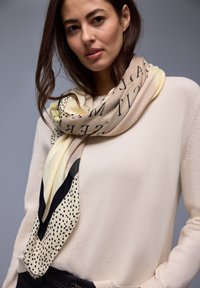 Jeune femme aux cheveux bruns portant un pull beige et une écharpe jaune-beige avec un texte noir et un motif à pois autour du cou.