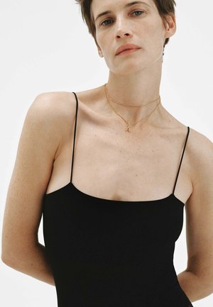 Mujer con cabello corto que lleva una blusa negra de tirantes finos y un collar dorado con un colgante en forma de "A" sobre un fondo blanco liso.