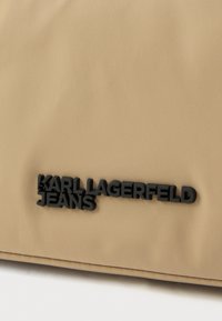 Bézs színű textil táska sima felülettel, amelyen egy fekete gumi logó található, rajta jól láthatóan a "KARL LAGERFELD JEANS" felirat.