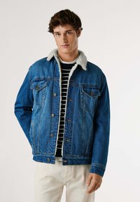 Jeansjacka i medelblå färg med fårskinnskrage, knäppning framtill, två bröstfickor och sydda detaljer, bärs över en randig skjorta.