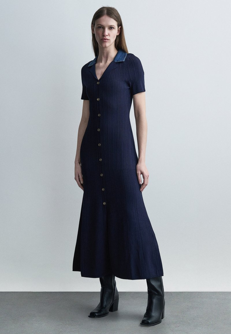 Γυναίκα φοράει φορεματάκι midi σε μπλε ναυτικό με ριμπ ύφανση, κουμπωτό, με γιακά από τζιν και μαύρα μποτάκια με τακούνι, στέκεται μπροστά σε απλό φόντο.