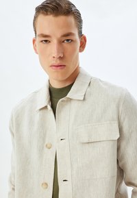 Lichtbeige linnen button-up jas met een enkele borstzak, zichtbare stikdetails en een relaxte pasvorm, gedragen over een donkergroen shirt.