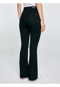 Sieviete melnās augsta vidukļa zvīņveida džinsās stāv, viena roka brīvi pie sāniem, redzamas baltas sporta kurpes.