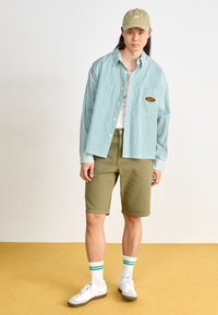Striped green and white button-up shirt, olive shorts, white sneakers, green cap, and striped white socks with green accents.