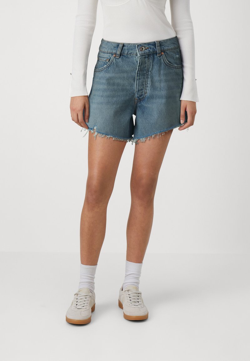 Lyseblå denimshorts med slitt søm, fem-lommers design og midt liv, kombinert med hvite sneakers og sokker.