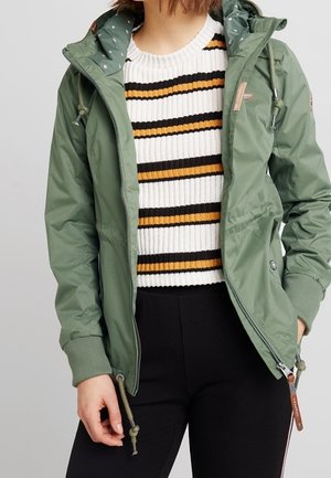 Person, die eine grüne Kapuzenjacke über einem weißen Pullover mit schwarzen und senffarbenen horizontalen Streifen trägt, kombiniert mit schwarzen Hosen.
