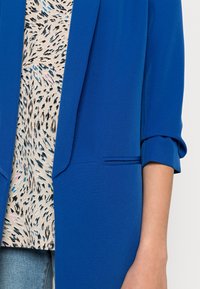 Blazer bleu roi à manches longues, doté d'une silhouette slim et d'une poche unique. En dessous, un haut beige avec un motif noir et bleu.