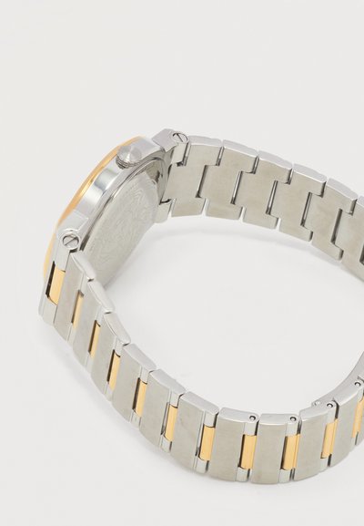 Náramok hodiniek zo spojov z nehrdzavejúcej ocele a zlatého tónu s okrúhlym zadným puzdrom hodiniek, bočnou korunkou a bezpečnou sponou, zobrazený na bielom pozadí.