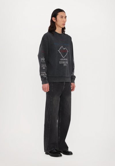Sweatshirt noir oversize avec des imprimés graphiques, assorti à un jean noir à jambes larges avec des coutures latérales contrastantes et des chaussures noires.