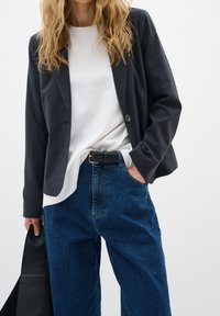 Tmavě šedý blazer přes bílou košili, doplněný volnými modrými džíny. Černý opasek a černá taška dotvářejí celý vzhled. Hladké textury, jednoduchý design.