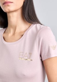 Gaiša rozā T-kreklu ar īsām piedurknēm, izgatavota no mīksta auduma, ar zelta kniedētā zīmola "EA7 Emporio Armani" uzrakstu priekšpusē un logotipa detaļu uz piedurknes.