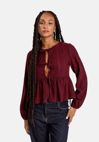 Blouse bordeaux à manches longues, à l'avant ouvert et avec des fermetures à nouer. Le tissu texturé présente des motifs complexes et un ourlet en peplum évasé.