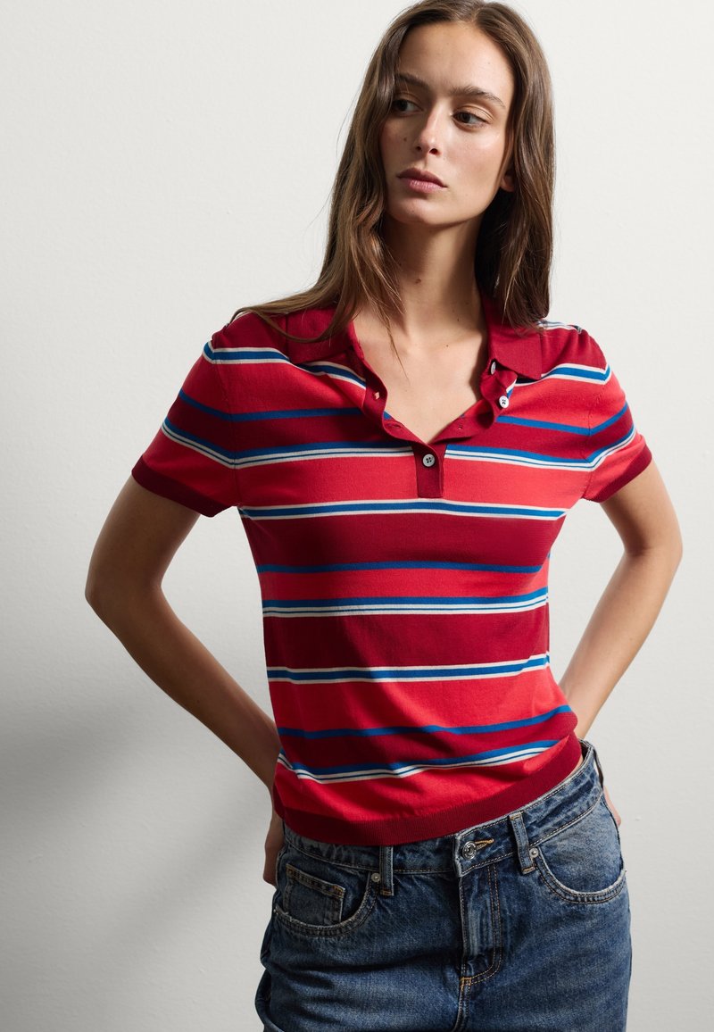 Rot gestreiftes Poloshirt mit blauen und weißen horizontalen Streifen, Knöpfen am Kragen und geripptem Saum, kombiniert mit blauen Jeans-Shorts.