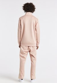 Světlé růžové teplákové soupravy z hladké tkaniny, s vysokým límcem a zlatými pruhy podél boků kalhot.