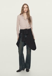 Hellviolettes langärmliges Hemd mit Kragen, kombiniert mit dunkel gewaschenen Schlagjeans. Ein schwarzes Stoffband, das in der Taille gebunden ist, sorgt für Textur und Kontrast.