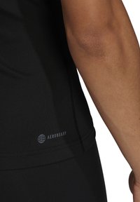 Gros plan sur le bras et le côté d’une personne portant un t-shirt de sport noir avec le texte et le logo « AEROREADY » près de l’ourlet.
