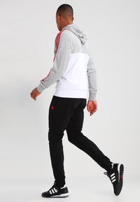 Sweatshirt à capuche gris et blanc avec des rayures latérales rouges, associé à un pantalon de jogging noir. Présente le logo Adidas et des chaussures de sport noires.