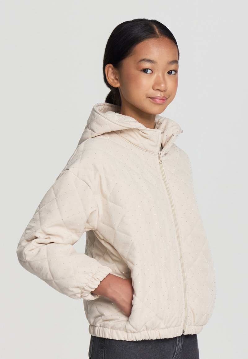 Giacca trapuntata beige con cappuccio, dotata di chiusura a zip, polsi elasticizzati e un sottile motivo luccicante. Tessuto morbido, design leggero.
