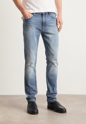 Homme portant un jean slim bleu clair avec délavé et petits détails usés, mocassins noirs et chemise blanche rentrée.
